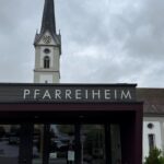 Eingang Pfarreiheim Schüpfheim mit rotem Plakat "Für bezahlbare Kitas" mit Würfeln, im Hintergrund Kirche Schüpfheim